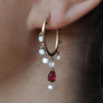 Close-up shot shows a woman's ear with gold earrings that feature a round cut diamond and a pear-cut ruby gemstone hanging