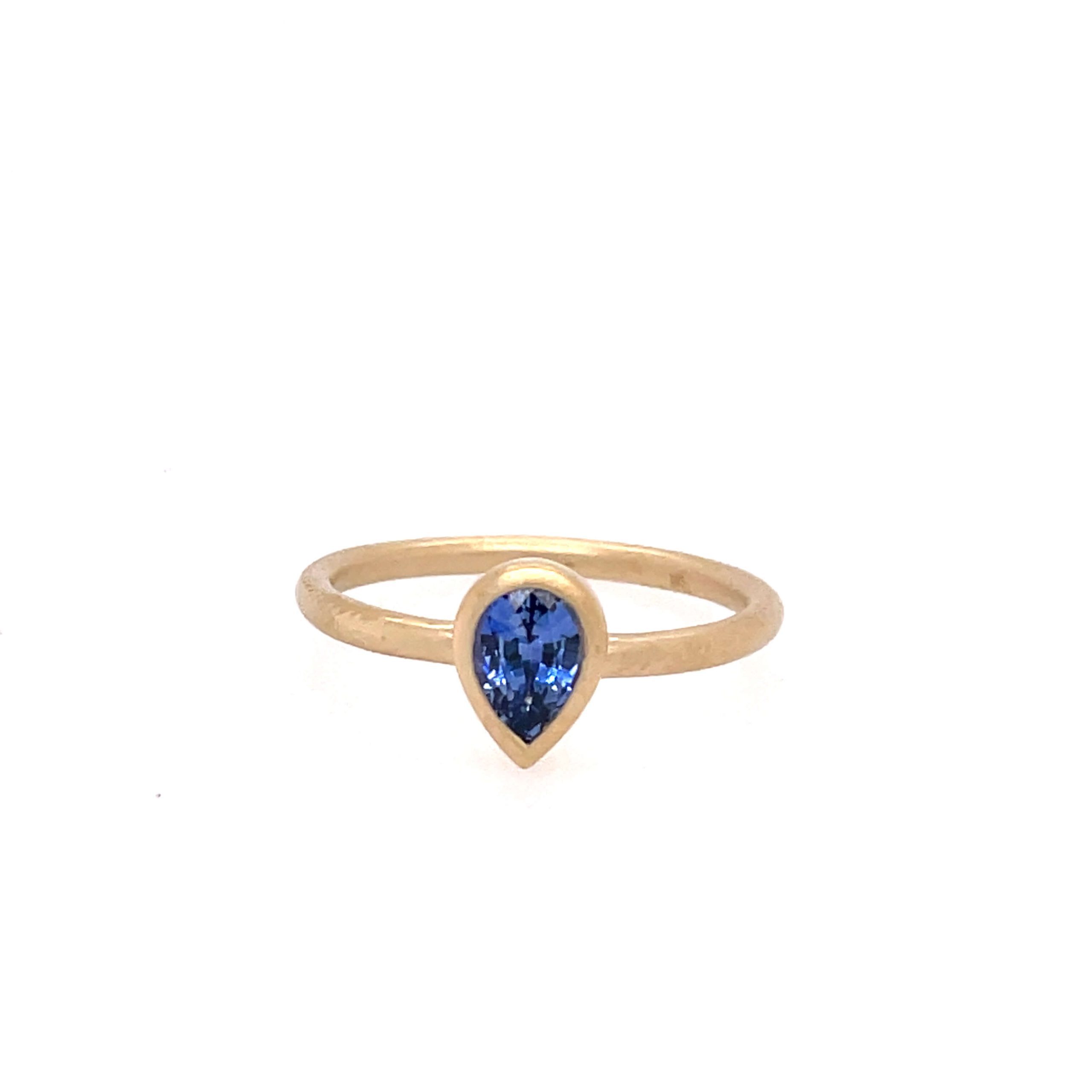 One yellow gold ring with a plain band and a bezel set pear shaped sapphire gemtone in the center, set against a white background.