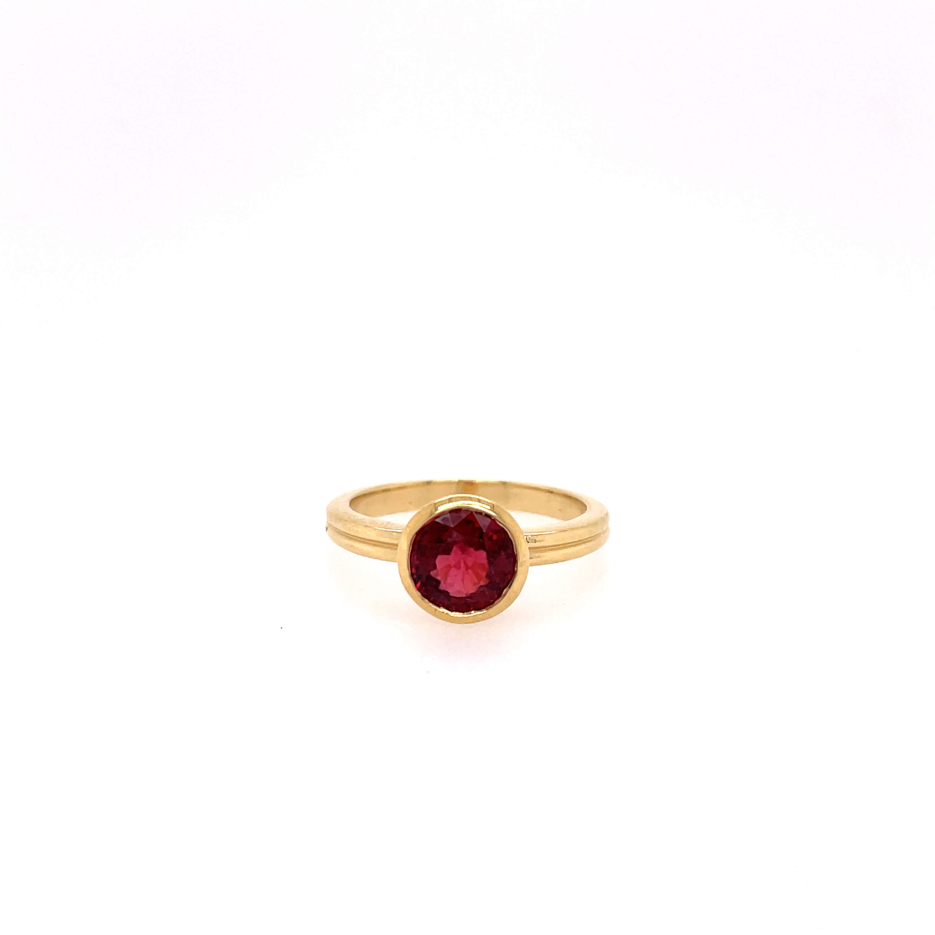One yellow gold ring with a plain band and a bezel set round shaped ruby gemtone in the center, set against a white background.