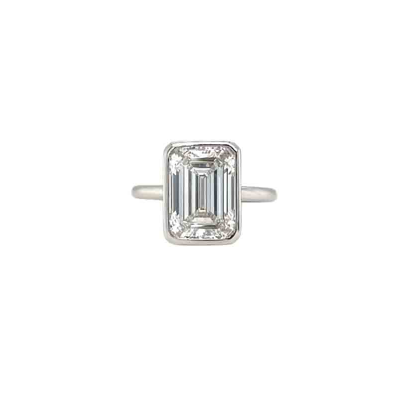 One white gold ring with a bezel set emerald cut diamond in a simple plain band. The ring rests in a white background.